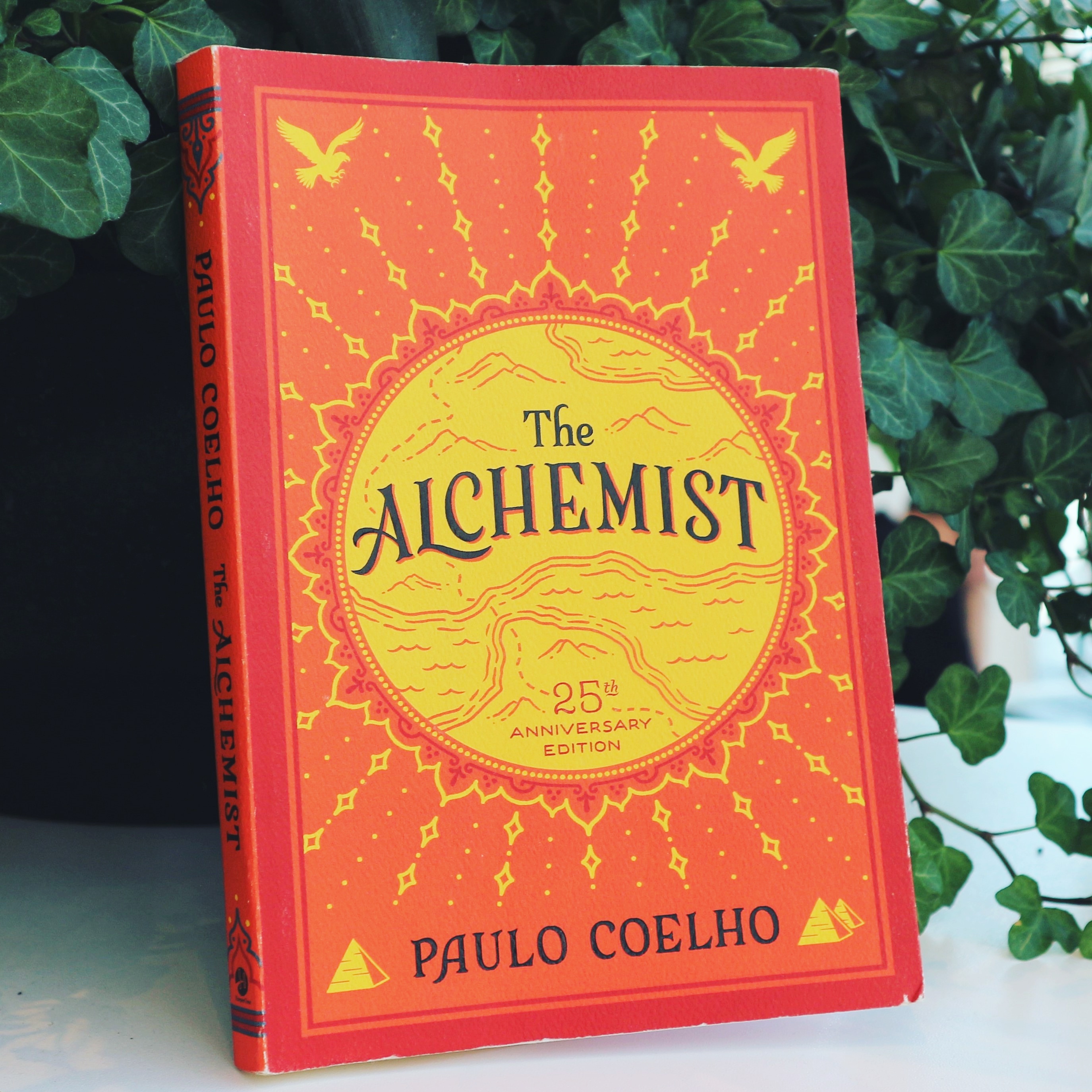 The Alchemist by Paulo Coelho cover. A red/orange colour with yellow map in the middle and yellow floral designs around.